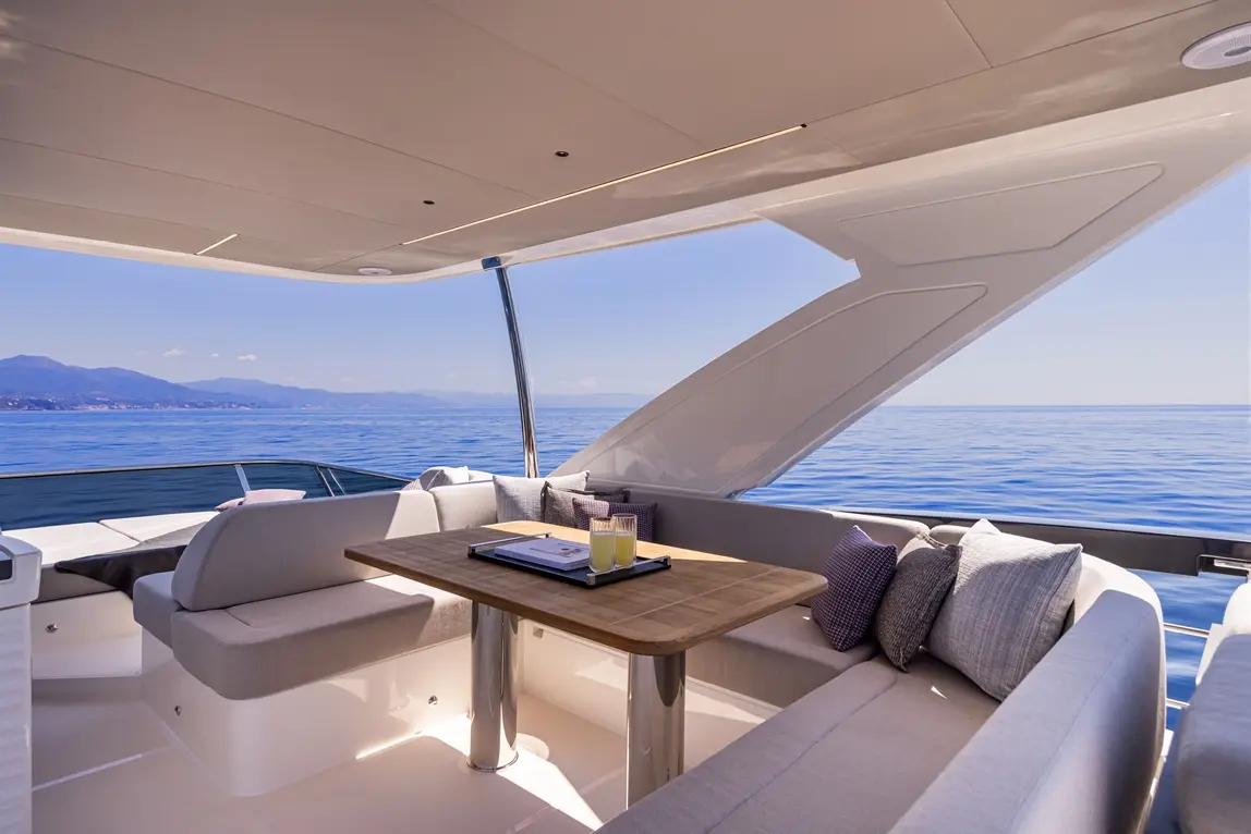 A stylish yacht interior features a wooden dining table, plush seating, and scenic ocean views under bright blue skies.